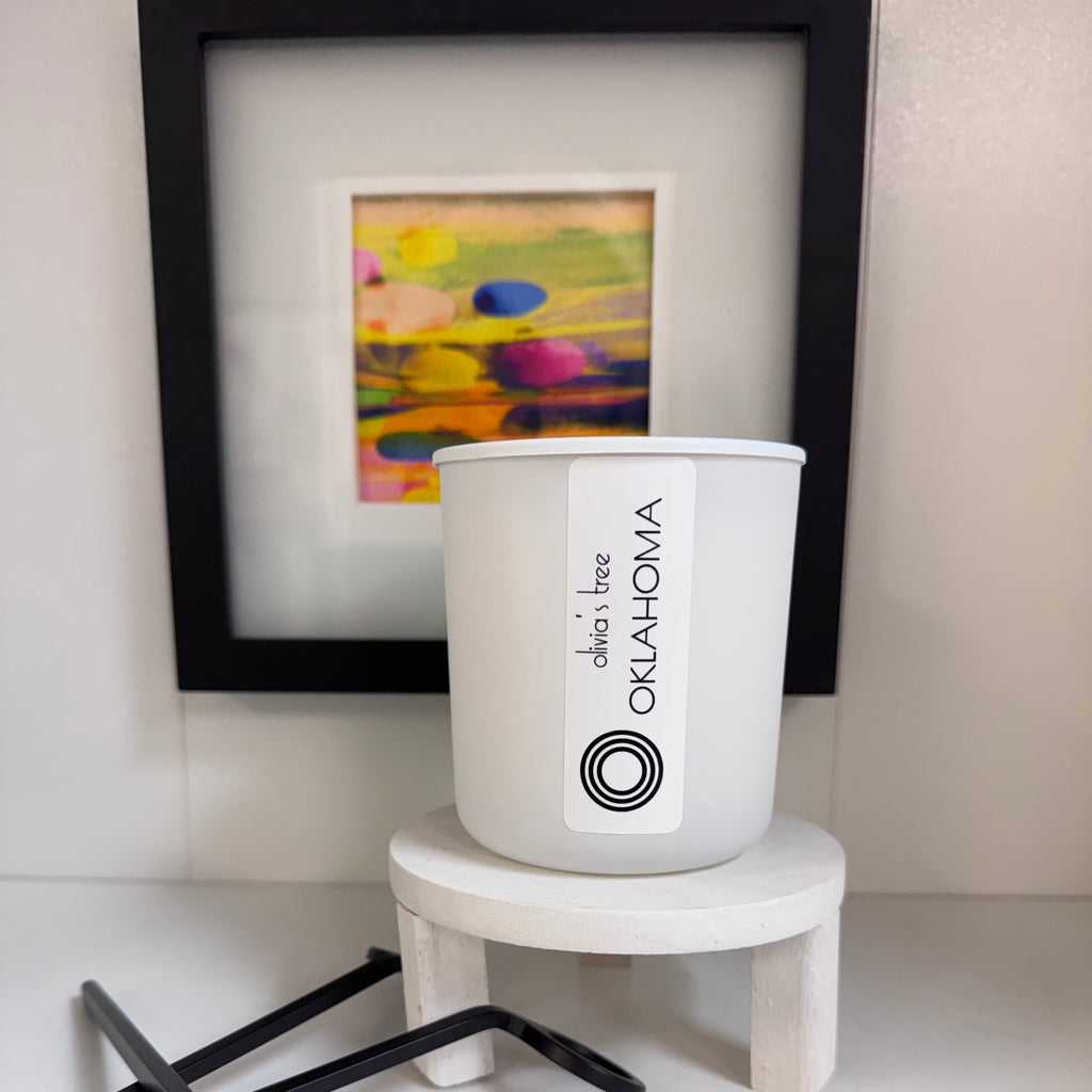 White candle with 'Oklahoma' label next to a framed picture on a white surface.